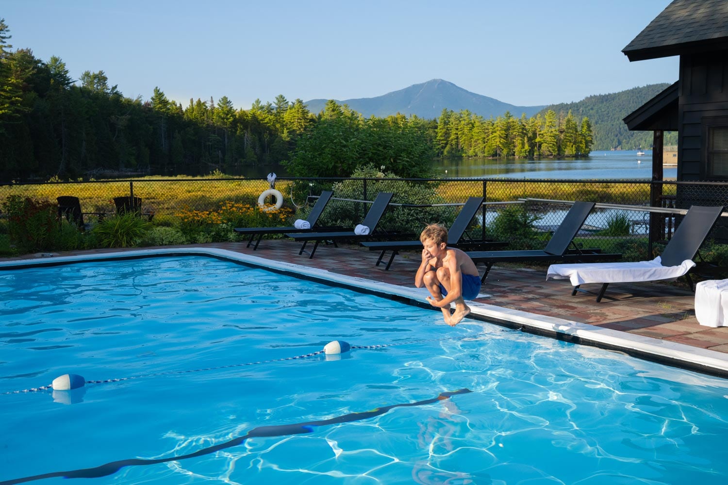 A man sits at the edge of an outdoor pool overlooking a lake and mountains.