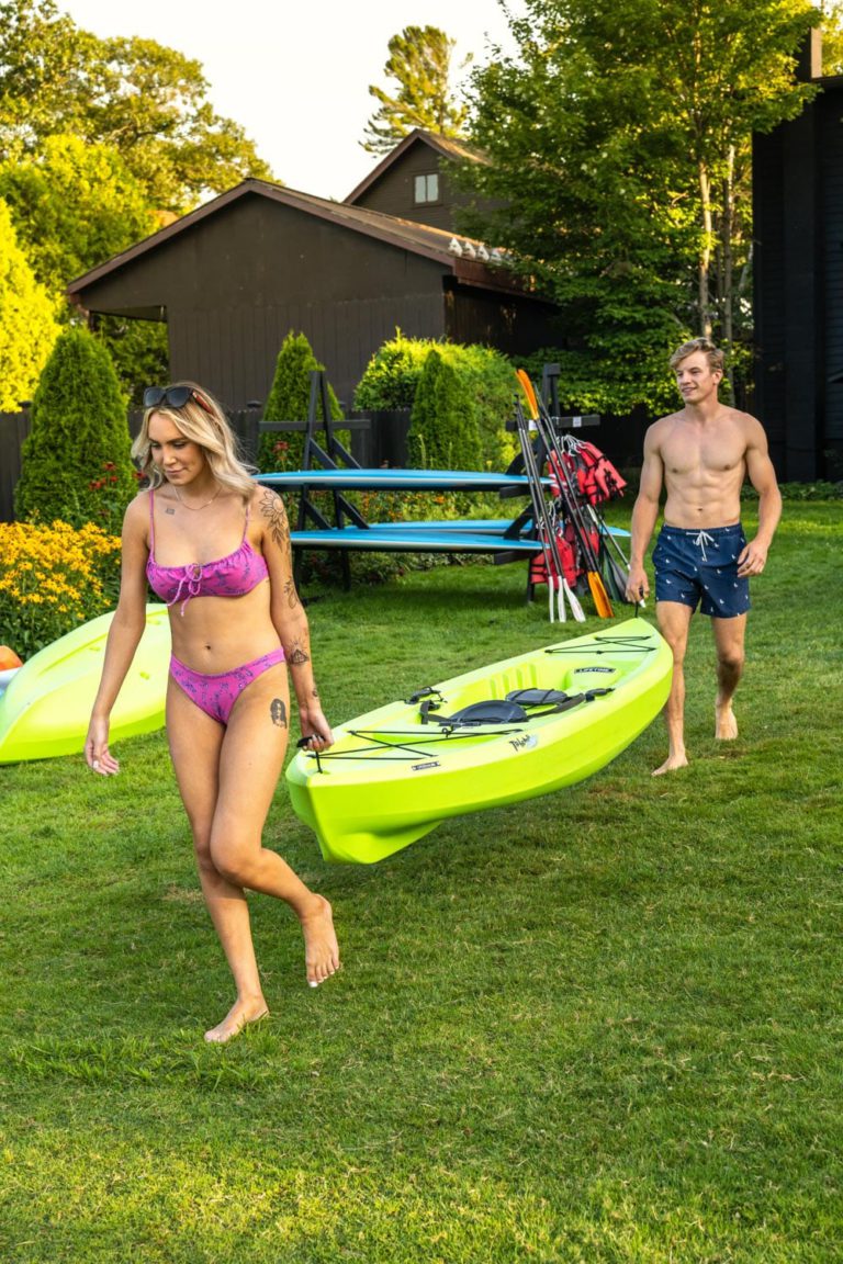 Two people in bikinis walking with kayaks in the yard.