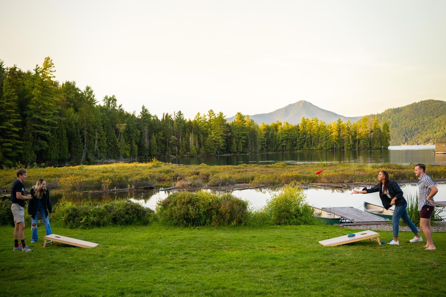 A group of people playing cornhole on a grassy area overlooking a lake with mountains in the background.