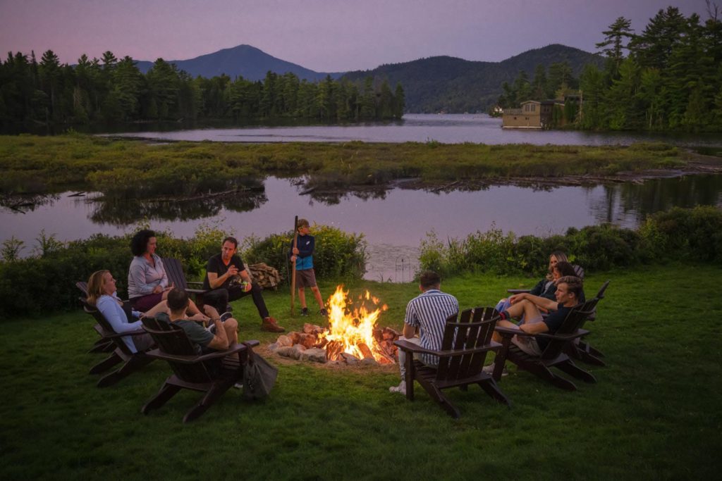 A group of people sitting around a fire pit in front of a lake.