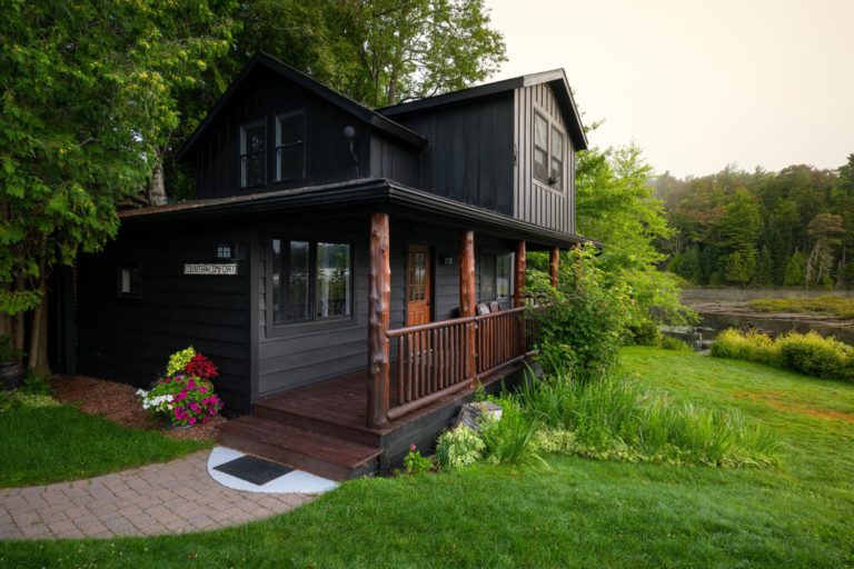A black cabin sits next to a wooded area.