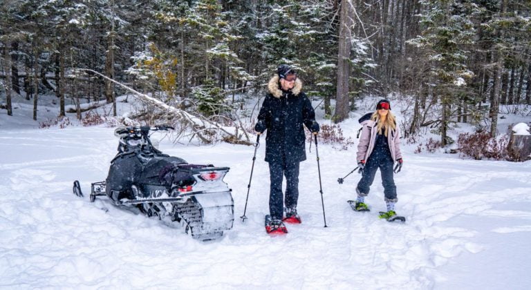 Two women standing next to a snowmobile in the woods.