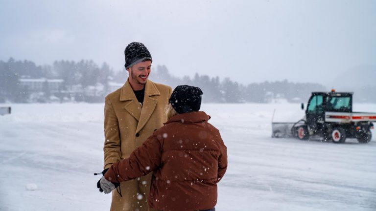 Two men standing on a frozen lake with a snow plow in the background.