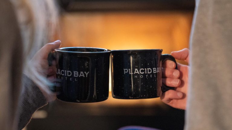 Two people holding coffee mugs in front of a fireplace.