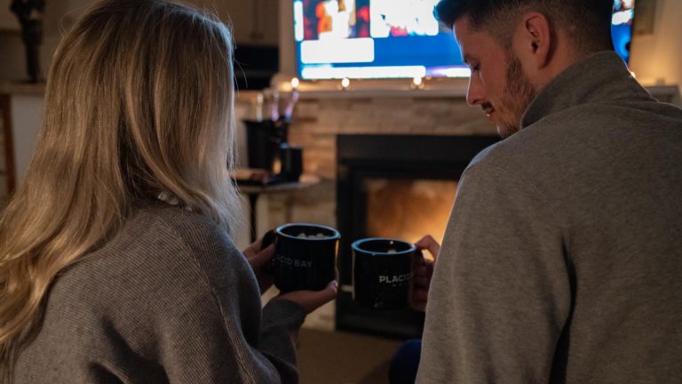 A man and woman holding coffee mugs in front of a fireplace.