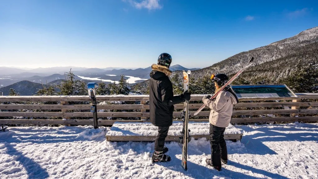 Two people standing at the top of a mountain with skis.