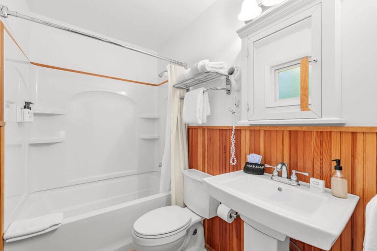 A small bathroom with a bathtub and shower, white fixtures, and wood paneling. A sink with a mirror above it is next to a toilet, and towels are neatly arranged.