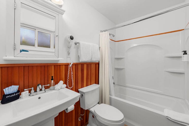 Bathroom with a white sink, toilet, and bathtub. Wooden paneling on walls, white towels, a mirror, and a wall-mounted hairdryer are visible.