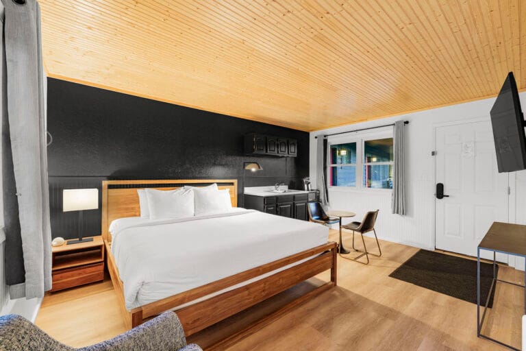 Cozy bedroom with a large bed, wooden ceiling, black accent wall, small kitchenette, table with chairs, and a wall-mounted TV.