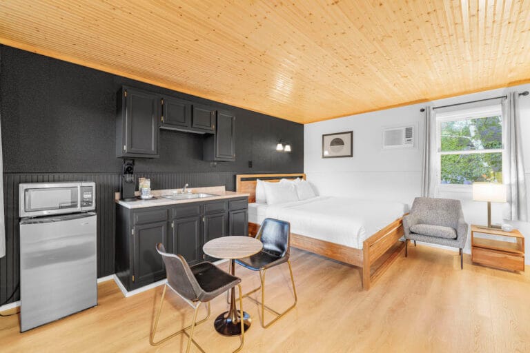 A modern hotel room with a wooden ceiling, featuring a bed, kitchenette with a black cabinet, microwave, mini fridge, small dining set, chair, bedside table with lamp, and a window.