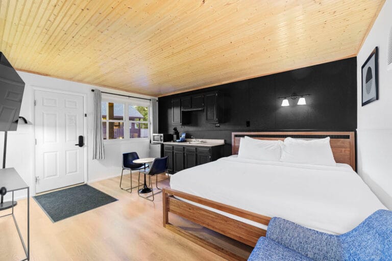 A modern room with a wooden ceiling, double bed, black kitchenette, and a small dining area. White walls, wall-mounted TV, and window with a view.