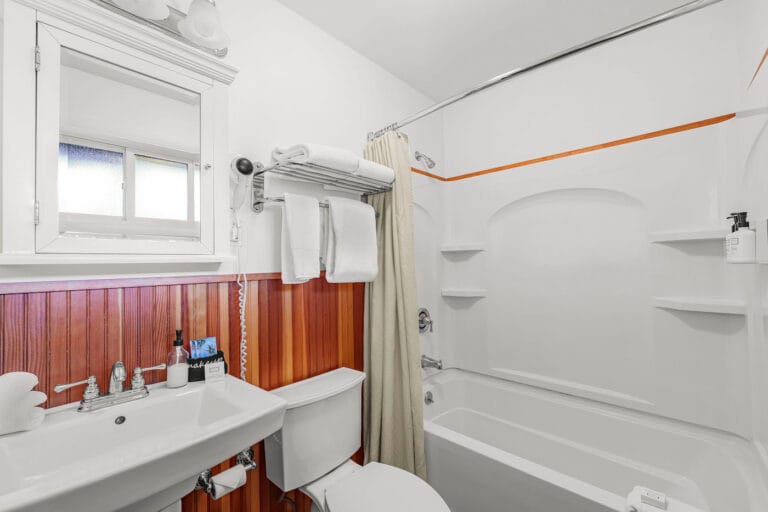 Bathroom with white walls, a bathtub with a curtain, a sink, mirror, and a toilet. Towels are neatly stacked on a rack above the toilet.