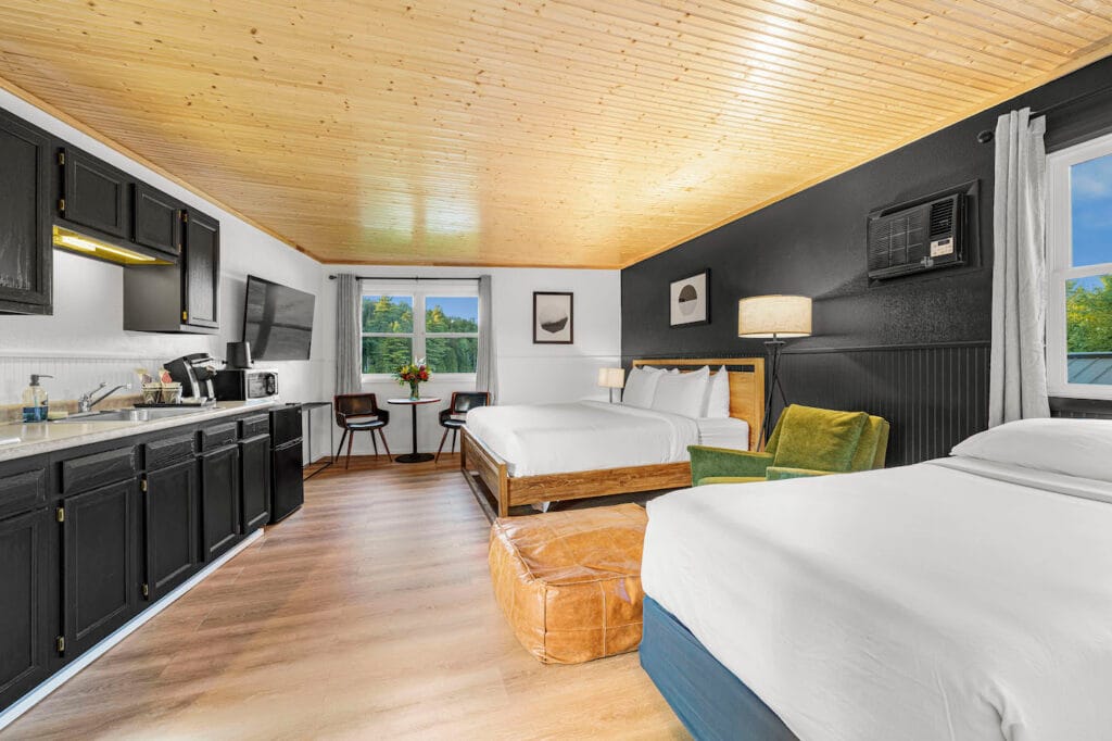 Hotel room with two beds, a kitchenette, TV, seating area with chairs and a table, large window, and wood-accented ceiling.