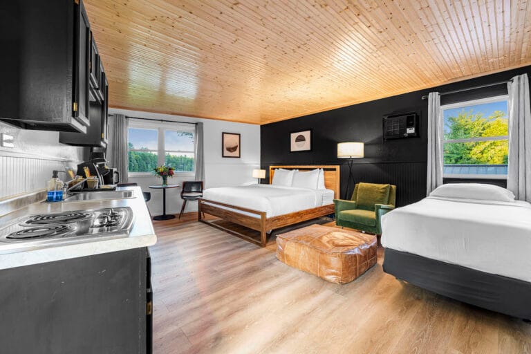 A cozy hotel room with two beds, a kitchenette, and a seating area. The room features wooden flooring, a light wood ceiling, and a window with a view of greenery.