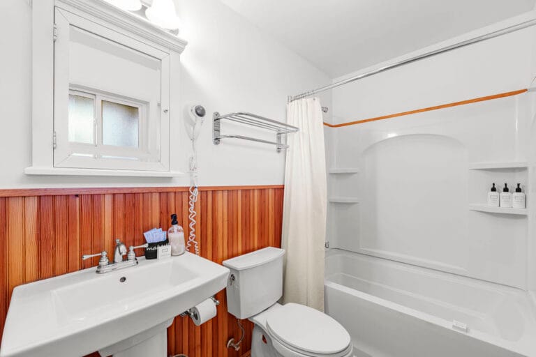 A bathroom with a white sink, toilet, and bathtub. The walls have white tiles and wood paneling. There's a mirror, a towel rack, and toiletries on the sink.