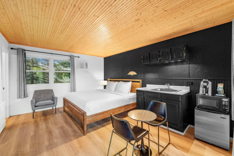 Bright hotel room with wooden ceiling, large bed, and kitchenette. Features chairs and a small table, window with curtains, and modern appliances.