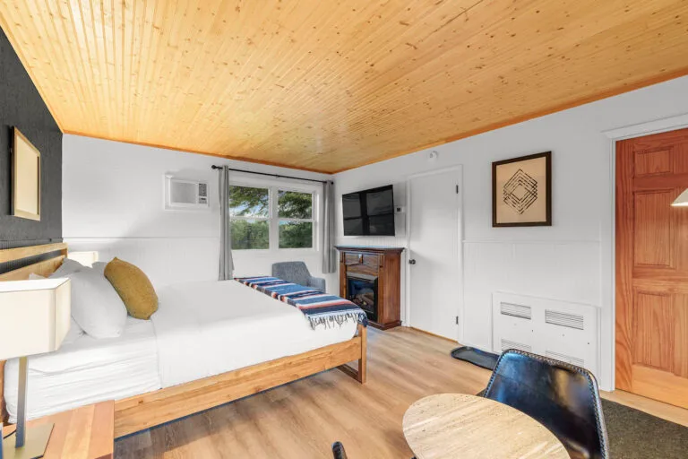 A cozy bedroom with a wooden ceiling, a bed with cushions, a wall-mounted TV, and a window with a forest view. A small table and chair are in the corner.