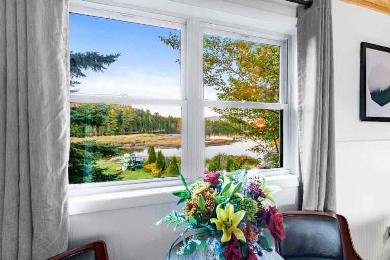 View through a window showing a scenic landscape with trees and a water body. Inside, a vase with colorful flowers is on a table by the window.