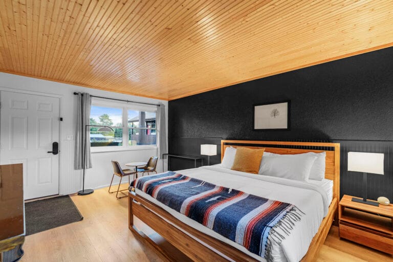 A cozy bedroom with a wooden ceiling, a double bed with a striped throw, two bedside lamps, a window, a small table with chairs, and a door. The walls are black and white.