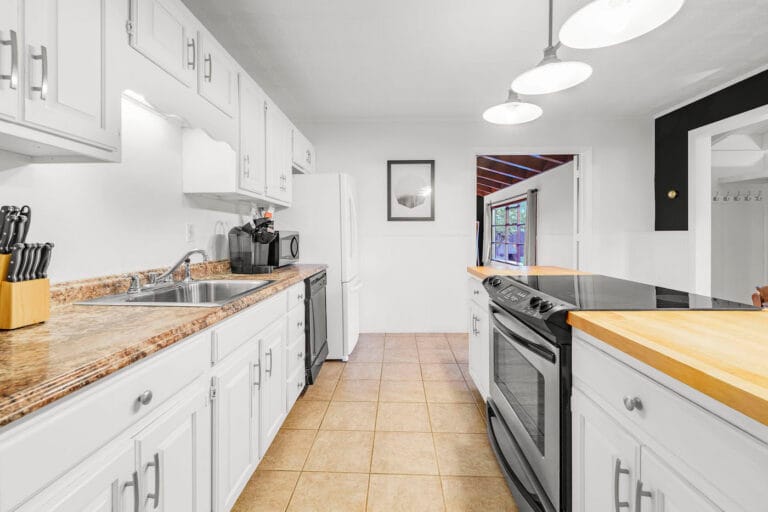 A bright kitchen with white cabinets, brown countertops, a stainless steel sink, black stove, and wooden chopping board. Three pendant lights hang from the ceiling. A framed picture is on the wall.