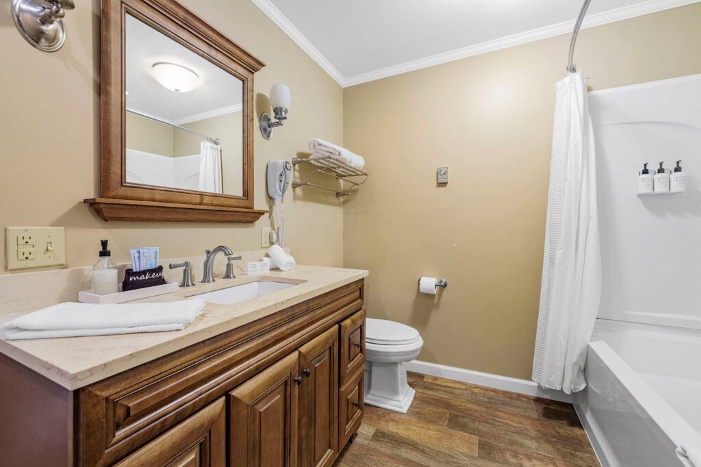 A bathroom with a wooden vanity, large mirror, sink, and toiletries. There's a toilet, towel rack, and a bathtub with a curtain. The floor is wooden, and walls are beige.