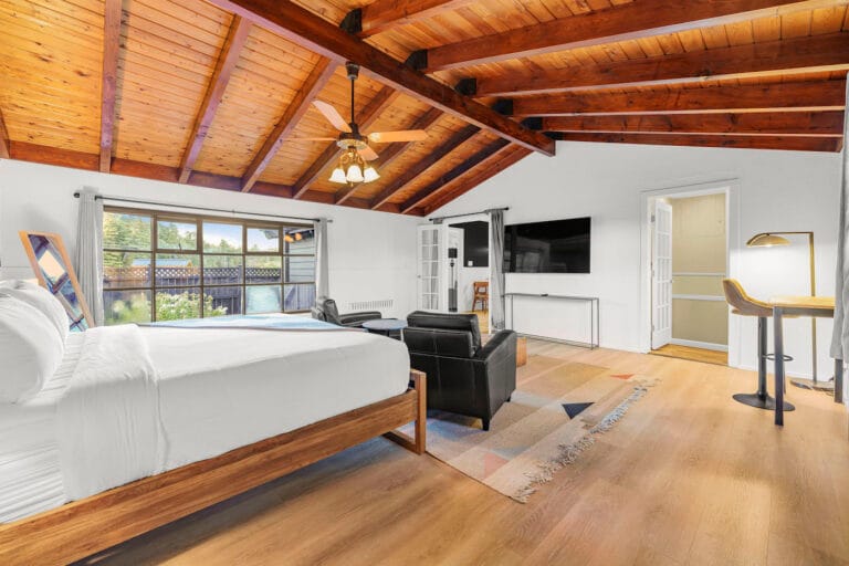 A spacious bedroom with wooden beams, a large bed, armchairs, a desk, and a wall-mounted TV. Large windows provide natural light.