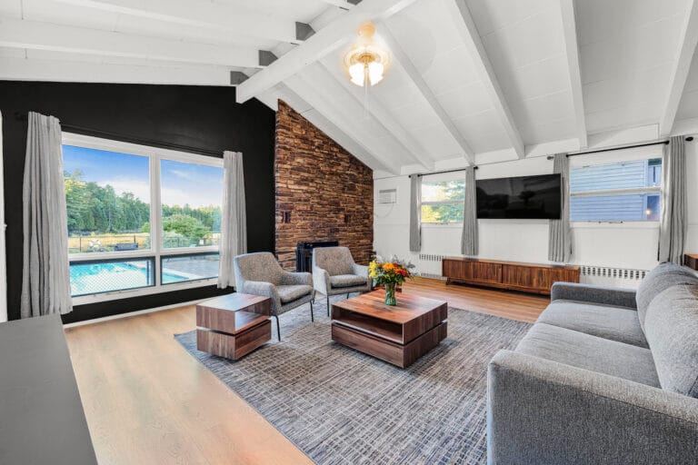 A modern living room with a vaulted ceiling, gray sofa, two chairs, a coffee table with flowers, a wall-mounted TV, and large windows overlooking a pool and trees.