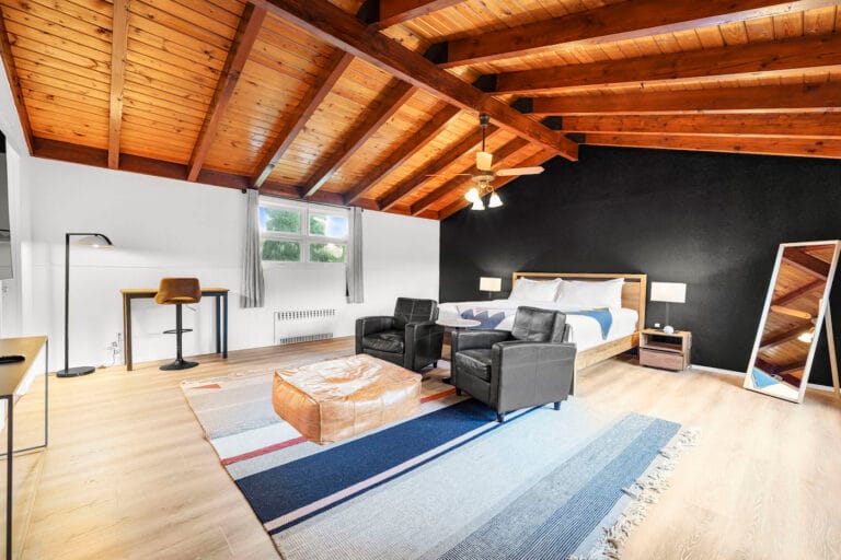 A spacious bedroom features a wooden ceiling, a bed, two armchairs, a large rug, and a floor lamp. A desk and chair are placed by the window. A free-standing mirror is on the right.