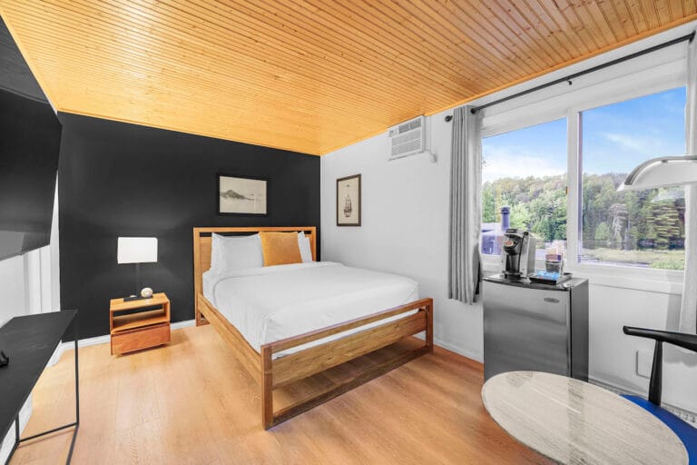 A modern bedroom with a wooden ceiling and floor, featuring a double bed, nightstand with a lamp, small fridge, coffee maker, and a window with a view of trees outside.