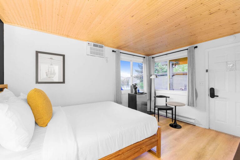 A modern bedroom with a wooden ceiling, white bed, and a yellow pillow. Two windows provide natural light, and a small table with chairs is near the door.