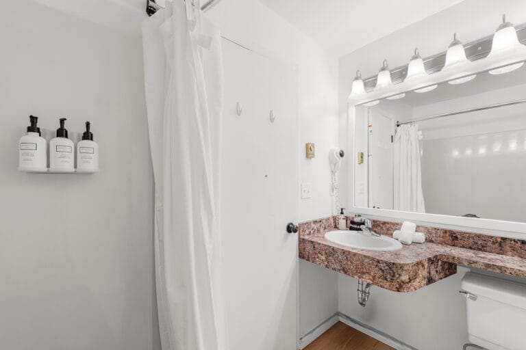 A bathroom with a white shower curtain, wall-mounted soap dispensers, a granite countertop sink, a large mirror with lights, and a wall-mounted hairdryer.