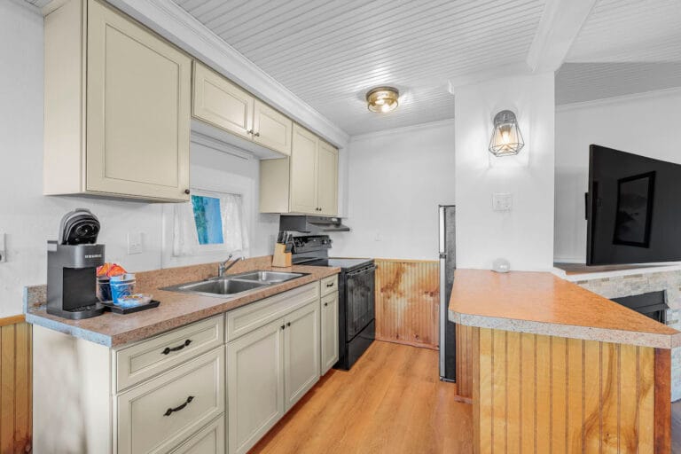 A kitchen with light cabinets, a black stove, and a countertop. A coffee maker and a basket of snacks are on the counter. Wood paneling and a wall-mounted TV are visible.