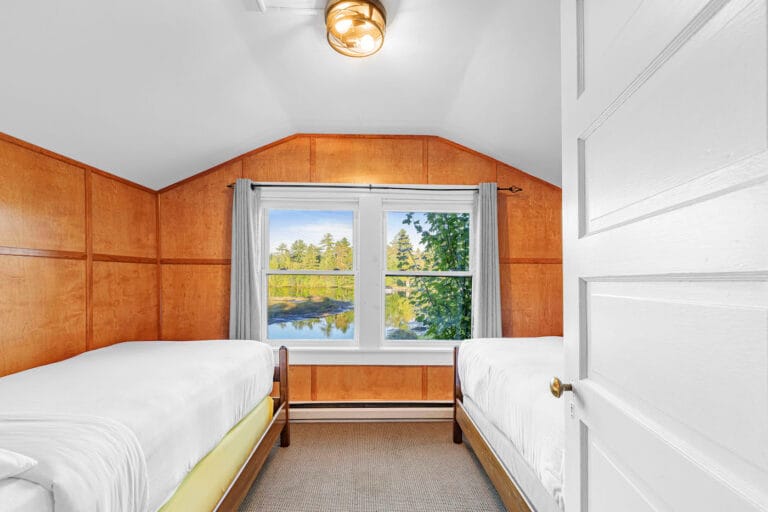 A bedroom with two single beds, wooden walls, and a window showing a view of trees and water.