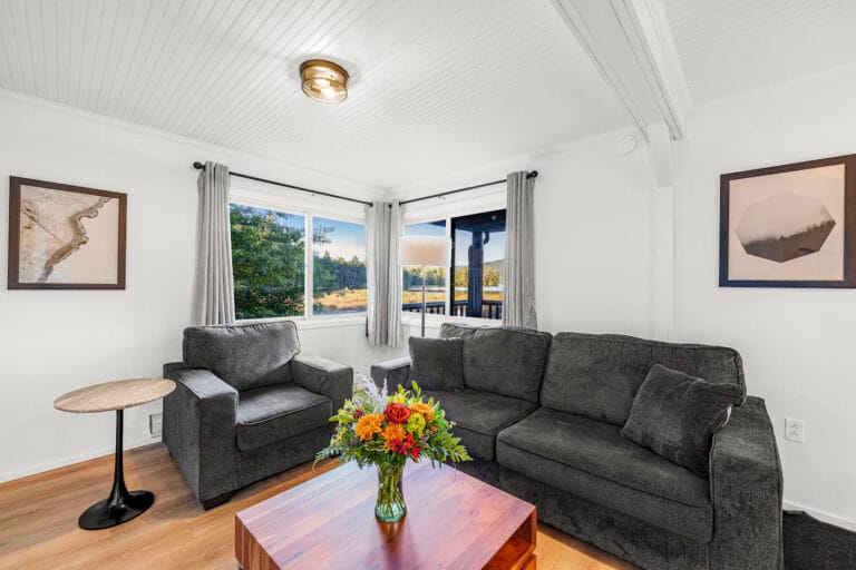 A cozy living room with two gray sofas, a wooden coffee table with a bouquet, framed art on the walls, and large windows displaying a scenic outdoor view.