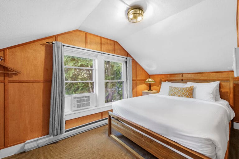 A cozy attic bedroom with wood-panel walls, a large double bed, white bedding, a small lamp on the side table, and a window with a view of greenery. An air conditioner is also visible.