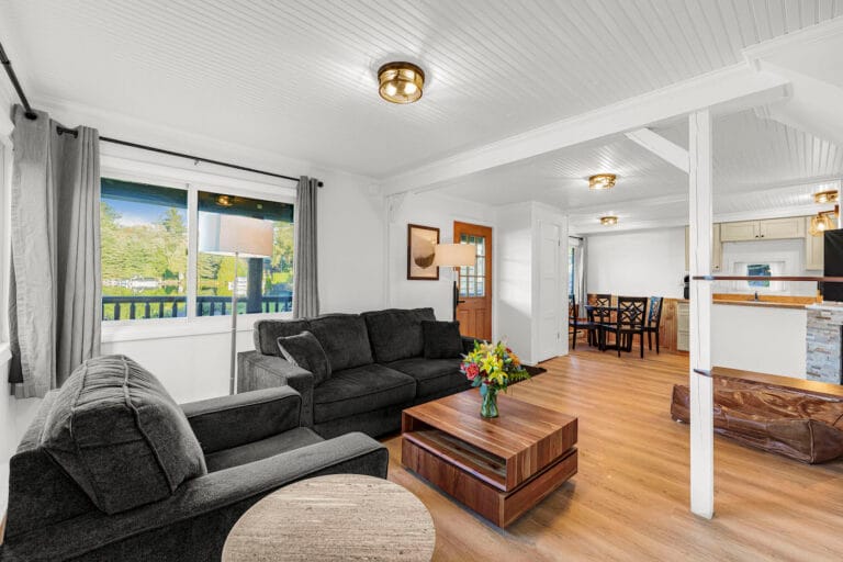 A living room with a dark sofa, a wooden coffee table with flowers, and a view of the kitchen and dining area. Large windows let in natural light.