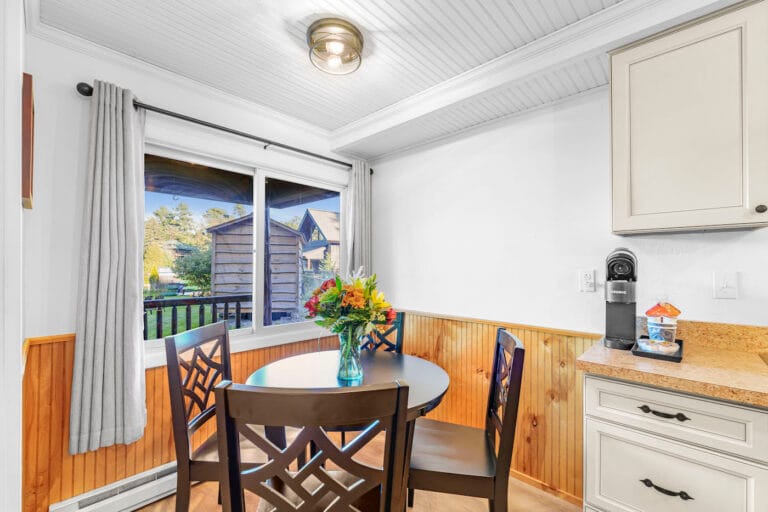 Cozy dining area with a round table and chairs, a vase of flowers, and a window view of a garden. Cream cabinets and a coffee machine are visible on the right.