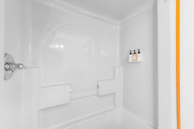 White bathtub with shower, featuring three mounted bottles of toiletries on a corner shelf, and an orange shower curtain partly visible on the right.