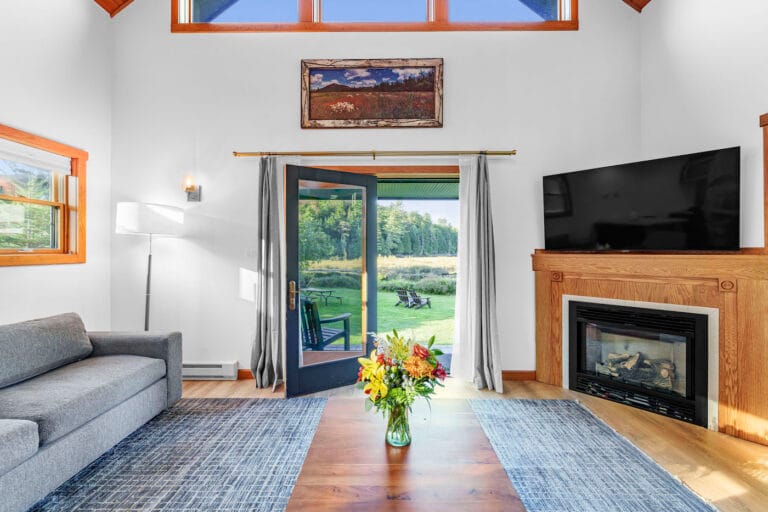 Living room with a gray sofa, wooden table with a vase of flowers, and a fireplace. French doors open to a grassy outdoor area.