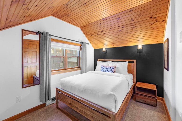 A cozy bedroom with a wooden ceiling features a large bed with white bedding, a dark accent wall, and wall-mounted lights. There's a wooden side table and a window with gray curtains.