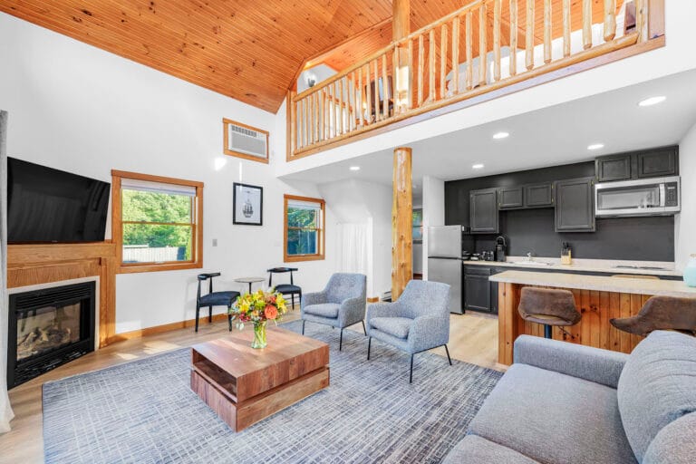 A modern living room with a loft features a gray sofa, armchairs, wooden coffee table with flowers, flat-screen TV, and kitchenette. The ceiling is wood-paneled, and large windows bring in natural light.