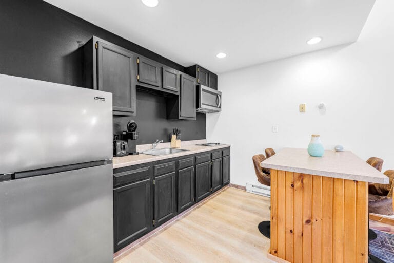 A modern kitchenette with black cabinets, a sink, a mini-fridge, and a small bar with two stools. Walls are white with black accents.