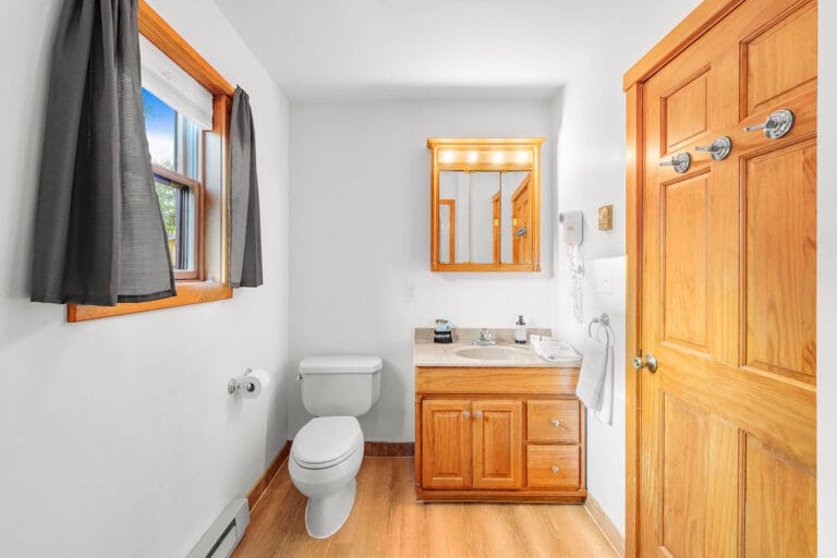 A bathroom with a wood vanity, mirror, toilet, and window with dark curtains. White walls, wooden door and floor.