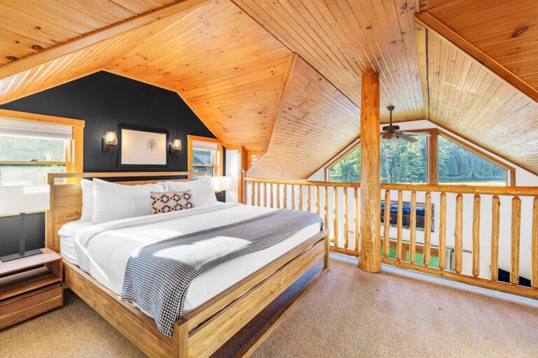 Loft bedroom with wooden ceiling, large bed, nightstands, and railing overlooking windows with forest view.