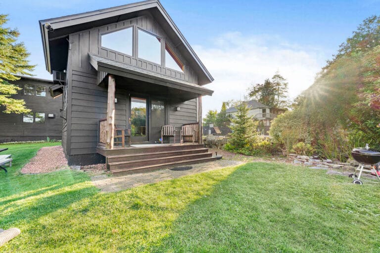 A two-story dark gray house with a wooden porch, grassy yard, and trees surrounding it on a sunny day. A small grill stands on the right side of the yard.