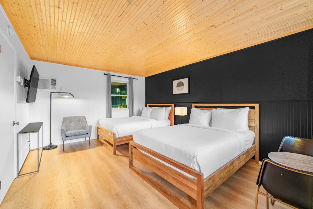 Modern hotel room with two double beds, wood flooring, black accent wall, wall-mounted TV, small chair, table with two chairs, and a window with a tree view.