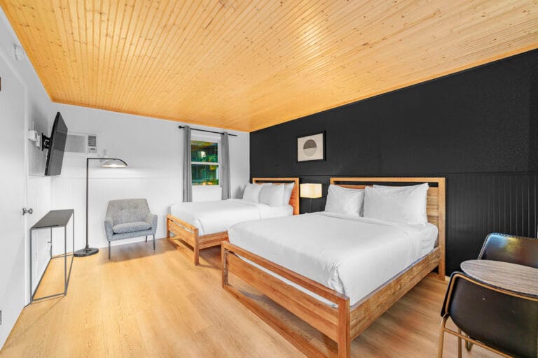 Modern hotel room with two double beds, wood flooring, black accent wall, wall-mounted TV, small chair, table with two chairs, and a window with a tree view.