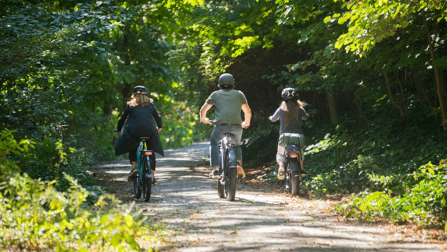 Three people ride bicycles on a shaded, tree-lined path during the day.