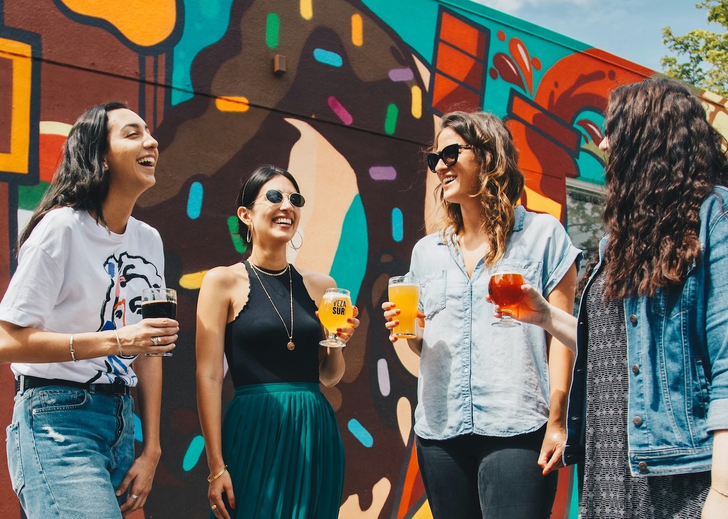 Four people stand outdoors in front of a colorful mural, holding drinks and smiling as they talk to each other.